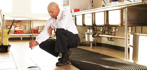 Inspector checking a floor drain in a commercial kitchen near stainless steel sinks