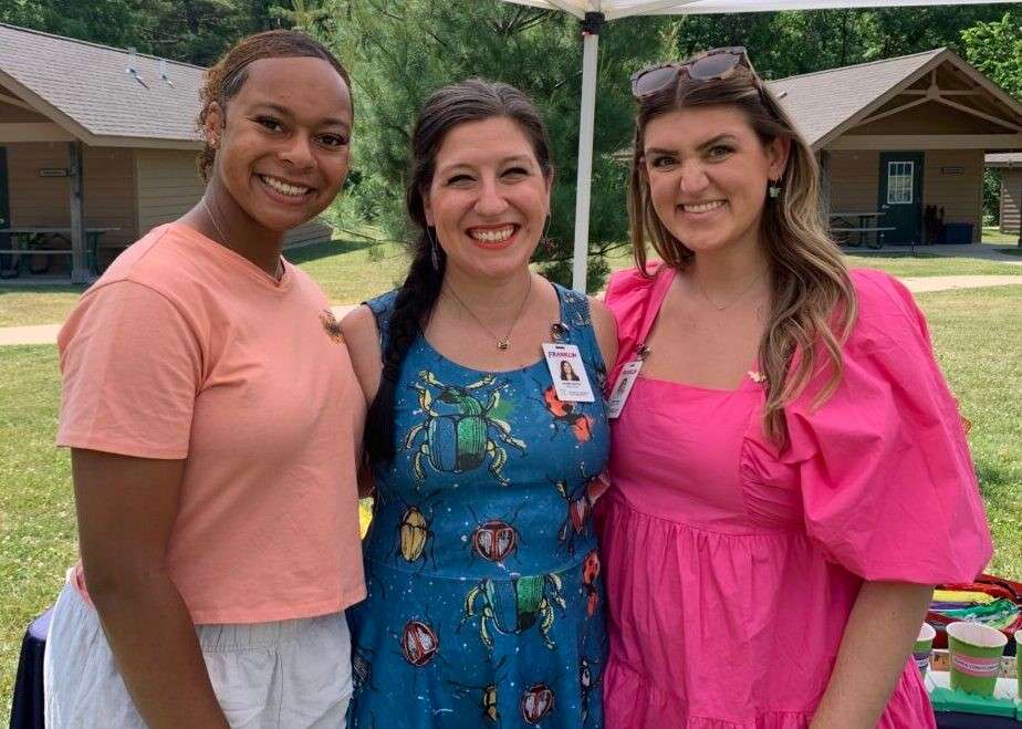 Three ladies posing together outdoors near cabins at a community event
