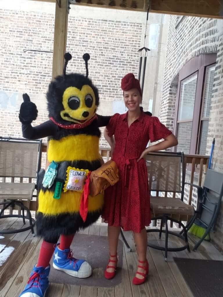 Person in a bee costume standing beside a person in a red dress on a wooden porch during a pollinator awareness event
