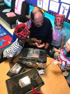 An adult sits at a table showing small animals to children wearing handmade hats in a classroom decorated with colorful posters and rugs—making it the perfect opportunity to visit your classroom and spark curiosity.
