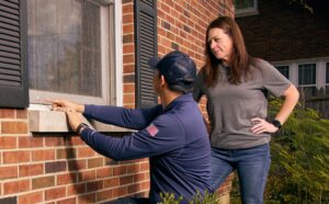 A man in a blue cap points at a window screen while a woman in a gray shirt stands next to him outside a brick house, possibly inspecting for signs of Odorous House Ant activity.