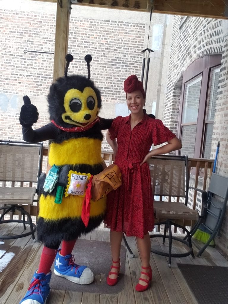 A person in a bee costume stands next to a smiling woman in a red dress and matching headwrap on a wooden porch, raising awareness for pollinator protection.