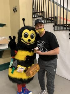 A person wearing a mask and casual clothes stands next to someone in a bee costume labeled "Flower," both smiling and posing for the camera indoors, raising awareness with their creative outfits.