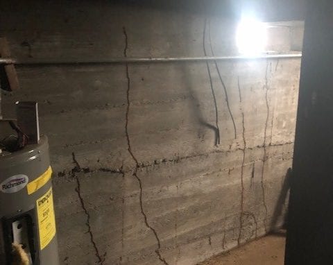 Dimly lit basement with a concrete wall showing multiple cracks, exposed pipes, a water heater, and a bare dirt floor—signs of ants and termites may be present in this vulnerable environment.