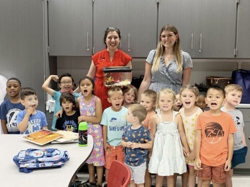 A group of young children and two adults stand together in a classroom, smiling at the camera; one adult holds a small aquarium tank, possibly sharing tips like how to get rid of wasps during their science lesson.