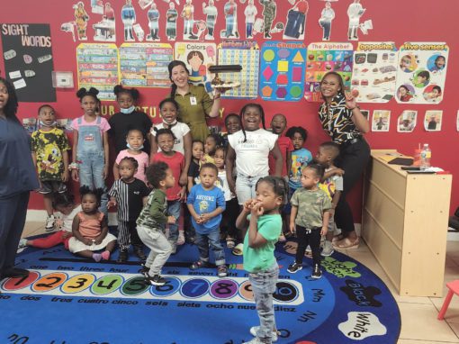 A group of young children and three adults pose and smile in a colorful classroom decorated with educational posters, a numbered rug, and a corner display about how to get rid of wasps.