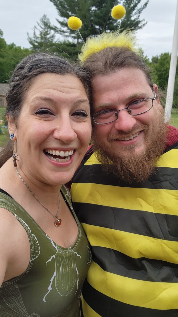Two people smiling for a selfie outdoors at a free STEM event; one wears a green dress with insect prints, the other is dressed in a bee costume with antennae. Trees and sky form the background.