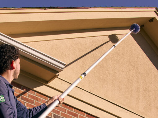 A person uses a long-handled brush to clean the exterior gable of a house under clear sky, maintaining cleanliness that's essential for effective commercial pest control.