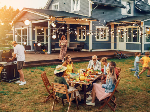 A group of people gather around a table with food in a backyard, while one person grills and children play near a large house decorated with string lights, occasionally swatting away fly problems that come with outdoor gatherings.