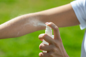 A person sprays a mist from a white bottle onto their forearm outdoors, with a green background.