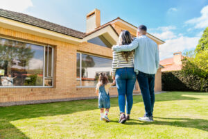 A man, woman, and young girl stand outside on the lawn, facing a brick house together with their backs to the camera.