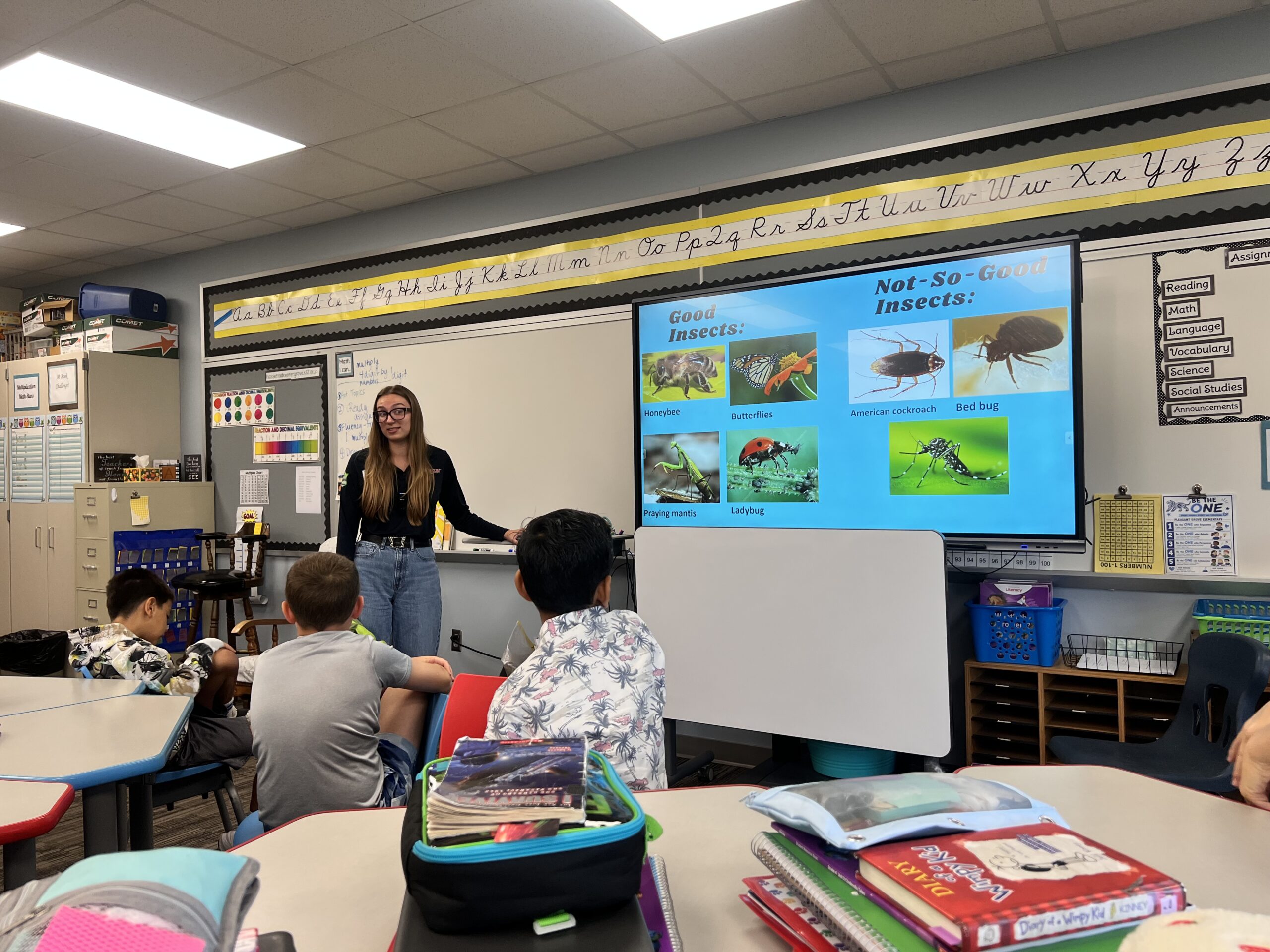 A teacher stands at the front of a classroom, presenting a slide about good and not-so-good insects to students seated at desks—introducing them to the fascinating world of “the bug squad.”.